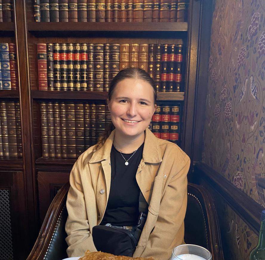 photo of a graduate student wearing a brown jacket in front of a bookcase