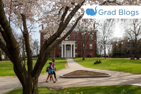 Image of two students walking on Tufts campus with Ballou Hall and a cherry blossom tree in the background